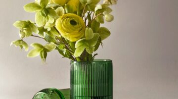 The NWTN Home Deco fluted emerald glass gravity pipe holding a vibrant bouquet of yellow ranunculus and green hellebore flowers, demonstrating its dual function as a tabletop vase.