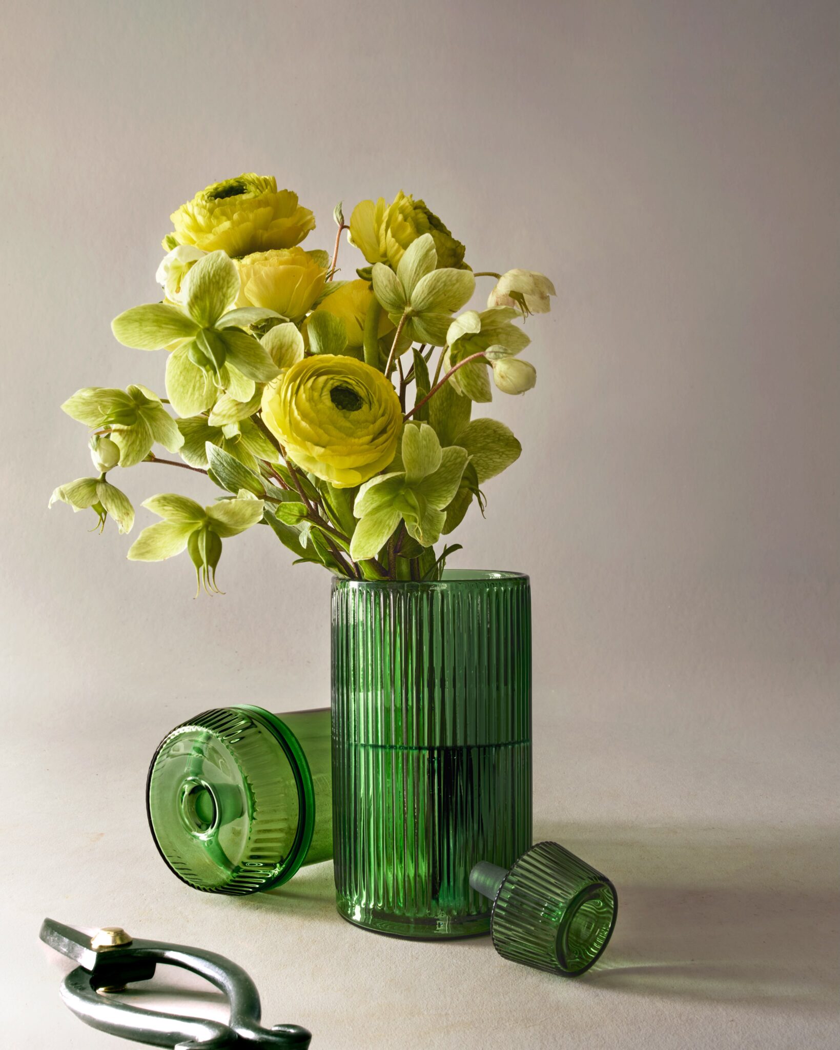 The NWTN Home Deco fluted emerald glass gravity pipe holding a vibrant bouquet of yellow ranunculus and green hellebore flowers, demonstrating its dual function as a tabletop vase.