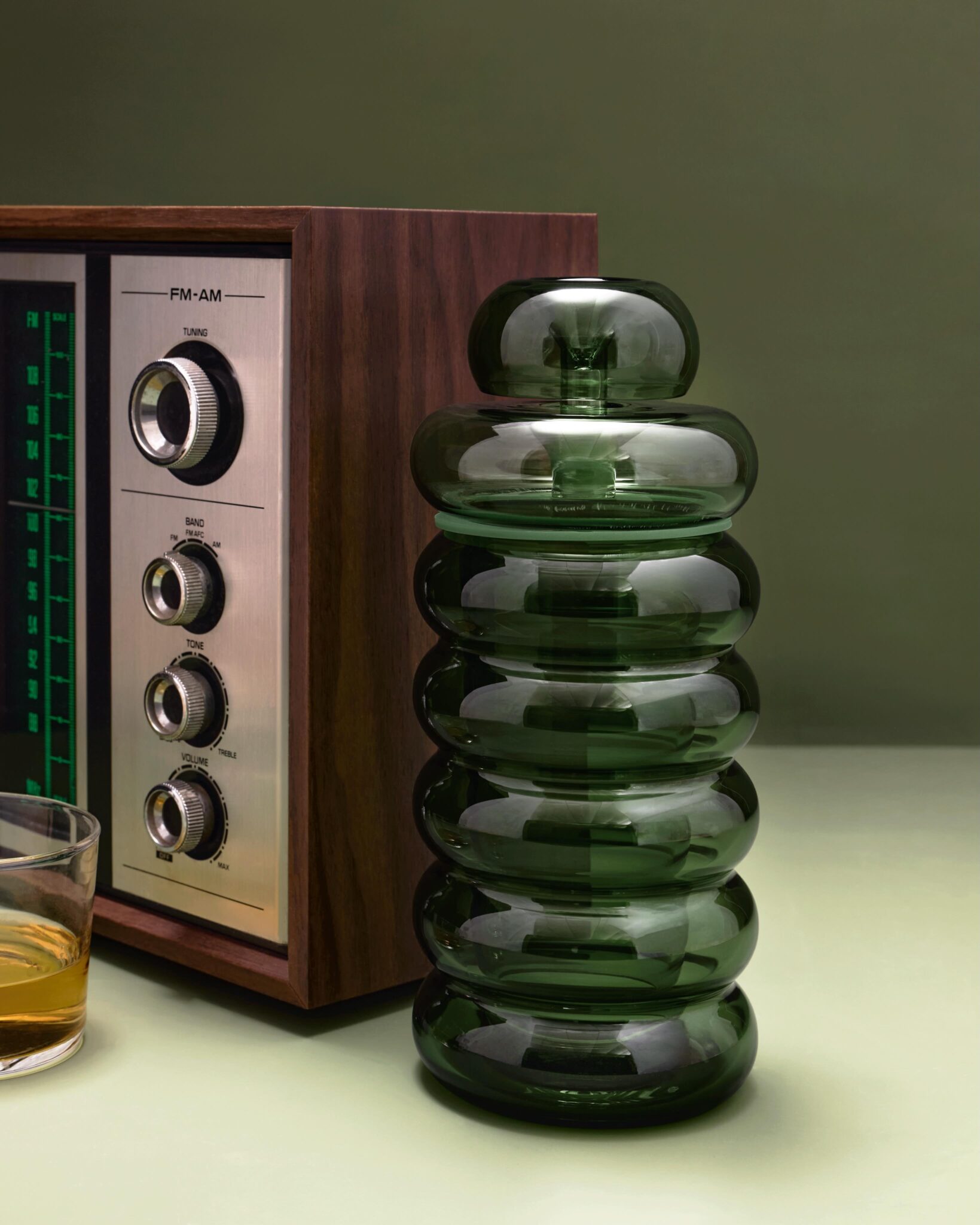 The NWTN Home Bolla Gravity Bong in emerald glass with its stacked bubble silhouette, displayed beside a vintage mid-century wood-paneled AM/FM radio and a glass of spirits.